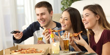 Three friends watching tv and eating pizza sitting on the floor in the living room in a house interior